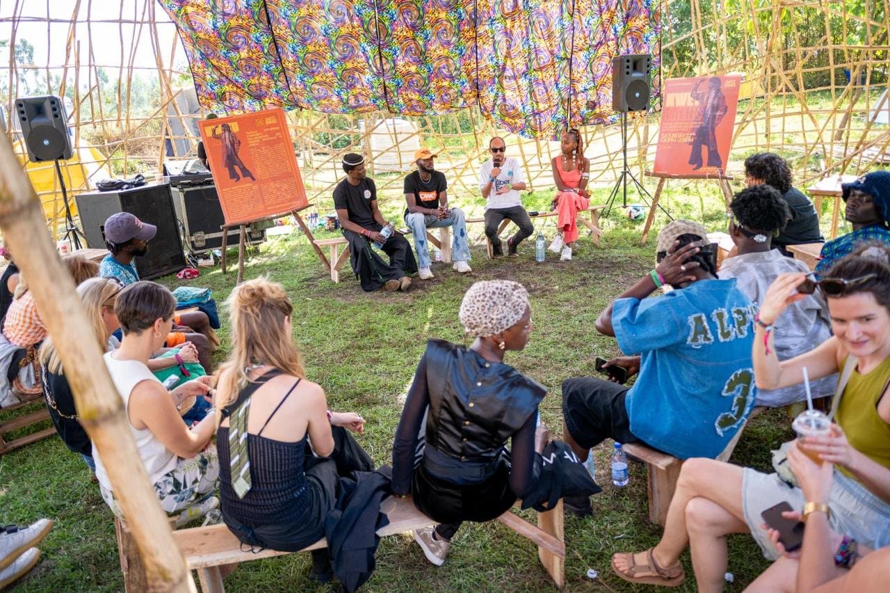 A seated audience listening to a panel beneath a patterned canopy in a Nyege Nyege promotional photograph.