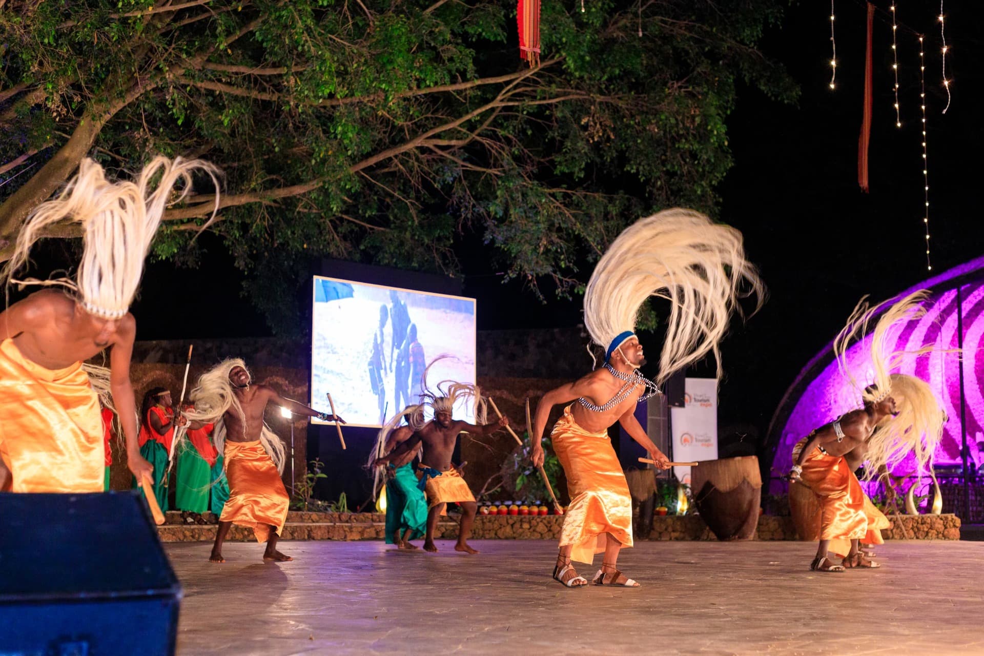 Ndere performers in orange wraps and tall headdresses dancing on an outdoor stage.