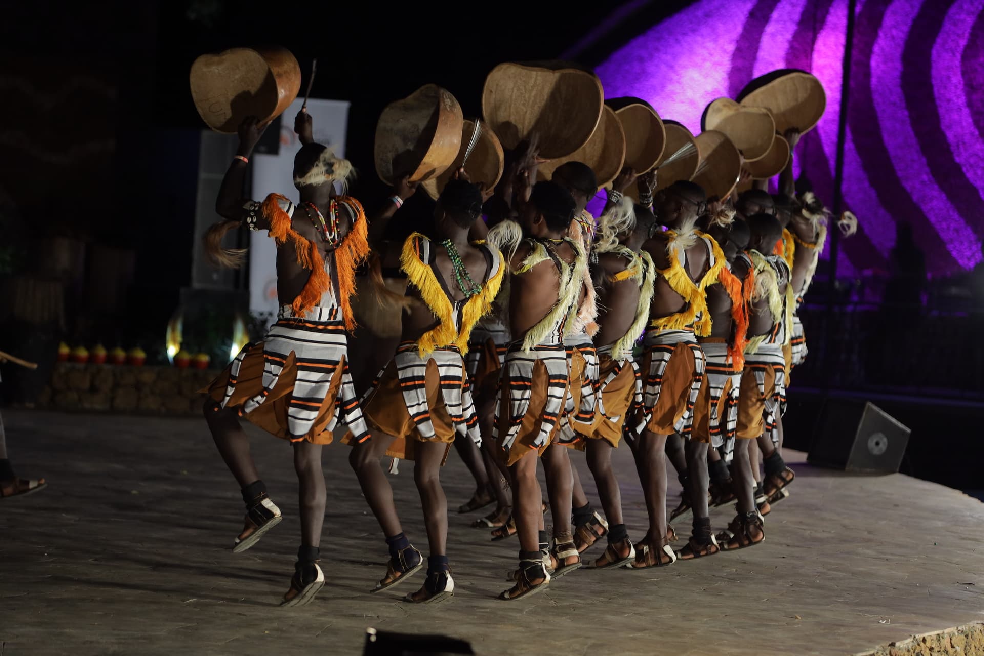 A line of Ndere dancers lifting calabashes above their heads during a stage performance.